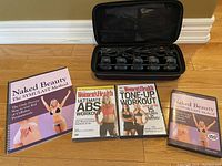Photo showing three exercise DVDs and a book titled 'Naked Beauty The SYMULAST Method' arranged on floor, alongside the IN Styler Topstyler hair curling system case closed.