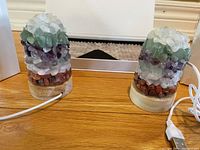 Two chakra healing crystal lamps on wooden bases with electrical cords visible, showing layered clusters of clear quartz, green, purple, and reddish stones.