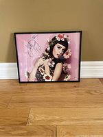 Framed signed photo of Katy Perry sitting on floor against beige wall. The size tipically 11.5x14.5 inches. The image shows the signature and floral theme with pink background.