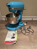 Front view of sky blue mixer with bowl and three attachments arranged in front