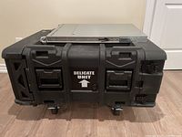 Front view of the closed black hard case, labeled 'DELICATE UNIT,' showing latches, handles, and wheels.