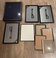 Top-down view of a set of nine frames on wooden floor showing a large 16x20 shadow box, three 11x17 frames, one 11x14 frame, and four 8x10 frames all with black borders and glass covers.