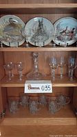 Photo showing three wildlife collector plates, crystal decanter, cut and etched crystal stems, and crystal rose mugs neatly arranged on wooden shelves.
