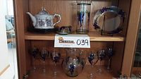 Front view of the glassware set on shelf including teapot, plates, wine glasses, pitcher, vase, and covered butter dish with purple and green grapevine handpainted design.