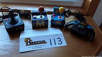 Four gaming controllers arranged on a windowsill showing Atari joystick, two Namco arcade controllers, and Batman controller.