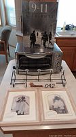Oster toaster oven with 9-11 Tribute coffee table book on top, black metal over door rack in front, and two matted and framed prints from Friends TV show placed at the front.