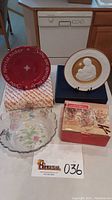 Photo of lot showing a red decorative plate with inscription and original box, Franklin Porcelain Silent Night 1975 plate on stand with box, glass serving bowl with floral pattern, and boxed set of four glass mugs