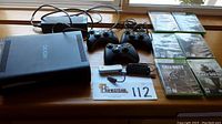 Xbox 360 console, power adapter, three wireless controllers, USB cable, and multiple Xbox 360 game cases arranged on a wooden surface near window light.