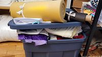 Bin packed with fabrics of different colors and textures, showing some rolled yellow foam on top and a variety of fabric pieces underneath.