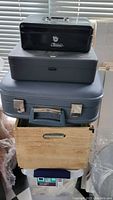 Stacked storage including black and gray cash boxes, blue vintage suitcase, wooden crate, and plastic bin below.
