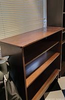 Dark wood wooden shelf unit, showing top surface and two open shelves below, positioned in front of a window with blinds.
