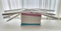 Stacked folded drapery panels in white with floral, pink and blue solid colors with long white paper blinds laying flat behind.