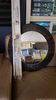 View of large round mirror with textured black frame leaning against a couch, alongside packaged Ikea white shelves and towel bars.