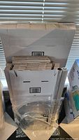 Photo showing stacked white corrugated pizza boxes, different sizes identifiable as 10" and 14". Several round cake boards visible in front, mostly white. Clear cellophane rolls also visible to the right.