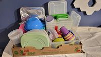 Plastic storage containers and kids cutlery and cups packed in a cardboard box.