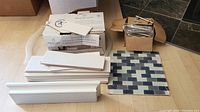Photo of stacked white ceramic wall tiles, a single sheet of blue-gray glass mosaic tiles, white floating shelf baseboard style pieces, a box of metal drawer pulls