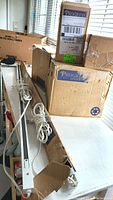 Two white 4-foot track lighting rails partially inside boxes with attached cords and plugs visible, set on a table by window.