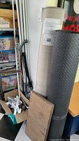 Photo of two large carpet or mat rolls, one gray patterned and one beige, standing upright with some flat boards and a vacuum cleaner visible.