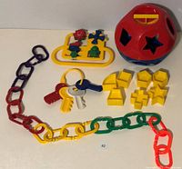 Top view of vintage baby toys including multicolored Boomerang Links chain, yellow spinner activity toy, colorful baby keys, and red and blue Tupperware shape sorter with yellow shapes.