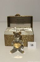 Glass teddy bear figurine holding a gold tone heart, placed in front of the cushioned fabric box with satin lining and 'Hugs and Kisses' inscription inside the lid.