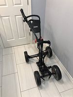 Side view photo of Cadie 4Runner golf push cart folded and standing upright against wall on tiled floor.