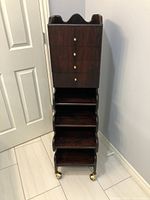 Front view showing the drawers with brass knobs above three stepped open shelves and brass casters at the base.