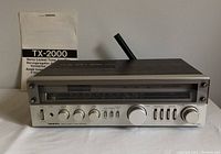 Front view of Onkyo TX-2000 tuner amplifier with instruction manual standing upright behind it on white surface.