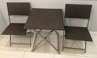Front view of the outdoor bistro set including the square table and two folding chairs with woven rattan-style tops in dark brown and silver metal frames.