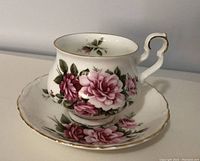 Porcelain cup and saucer with pink rose design and gold trim, shown from a front angle highlighting floral pattern and handle detail.