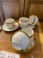 Five white Limoges china teacups and five matching saucers arranged on a wooden surface, displaying gold rim and decorative gold handle. Photograph shows top and side of cups with one cup tipped to display Limoges branding on the bottom.