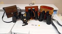 Overview of three pairs of binoculars, small microscope, and cases on white surface.