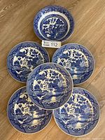 Five round blue willow patterned dinner plates arranged in a fan shape, showing scenic landscape and bird designs with visible wear and chips on edges.