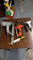 Overview of all three stapling tools - Paslode brad nailer, Campbell Hausfeld air stapler, Black & Decker electric stapler on a wooden table.