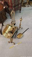 Photo showing brass fireplace tools on carpet including tool stand with tools, bucket, and two andirons with a striped chair in background.