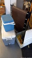 Photo of two cooler boxes stacked on a white table and a brown collapsible camp cot folded and leaning behind them
