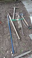 Full view of the 7 tools laid on ground showing the pry bar, axes, pick axes, spade, and peavey tied together.