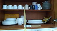 Dinnerware set inside cabinet on two shelves including stacked white plates, bowls, ceramic mugs of various colors, glass bowls