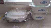Four Corning Ware ceramic cookware items with glass lids stacked and displayed on kitchen counter. White pieces with blue floral pattern visible on sides. One large 10-inch square casserole with metal rack and clear glass lid shown on left.