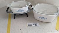 Photo showing both Corning Ware casserole dishes with their lids on white countertop. The 10-cup dish is on a black riser, the 48-ounce dish is beside it.