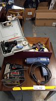 Full view of lot including Craftsman Auto Scroller Saw, heat gun, glue gun, soldering tools and accessories in a wooden and plastic container on floor with some cardboard boxes behind.