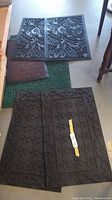 Five doormats arranged on the floor including two large black rubber mats with leaf and floral patterns, a small brown rubber mat, and two green nylon mats visible underneath.