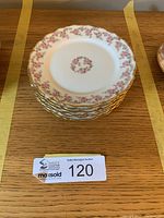 Stack of six white porcelain side plates with pink floral wreath pattern and gold trim on scalloped edges.