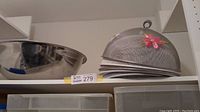 Metal bowls and wire mesh food covers stacked on a shelf, showing several large metal bowls and multiple dome-shaped mesh food covers.