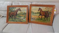 Two wood framed paint by numbers paintings displayed on a sofa, each depicting farm horses in natural settings with fields and fences in the background.