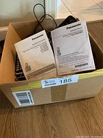 Box containing phones, manuals, and accessories from the lot. Visible are Panasonic user manuals labeled KX-TGDA20 and operating instructions.