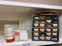 Close-up of sewing accessories including bobbins, string spool, tape measure, and package of Singer machine needles next to plastic containers.