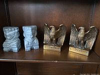 Two pairs of bookends displayed on a wooden shelf: a pair of gray carved stone bookends with a stepped design and a pair of metal eagle bookends with '1776' inscribed at the base.