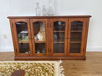 Front view of wooden sideboard cabinet with four glass panel doors, showing medium-brown wooden finish, visible shelving inside and two round knobs per door.