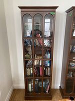 Front view of tall narrow wooden cabinet with glass-paned door containing multiple shelves.