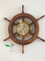 Front view of wooden ship's wheel clock with brass housing mounted on a white wall.